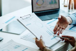 Zwei Personen im Anschnitt vor Schreibtisch mit Laptop und Papieren. 