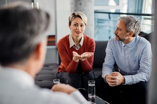 Ein weißes Paar mittleren Alters sitzt nebeneinander auf einer grauen Couch. Die Frau hat kurzes blondes Haar und spricht mit nach oben gerichteten Handflächen, ihr Partner hat kurzes graues Haar und sitzt mit verschränkten Händen, seine Körpersprache zeigt, dass er ihr zugewandt ist und aufmerksam zuhört. Eine Person mit kurzen grauen Haaren, vermutlich ein Therapeut, sitzt dem Paar gegenüber. Auf dem Tisch vor dem Paar stehen zwei Gläser mit Wasser. 