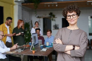Eine Person mit Brille steht rechts im Vordergrund mit verschränkten Armen, im Hintergrund steht eine Gruppe an einem Tisch